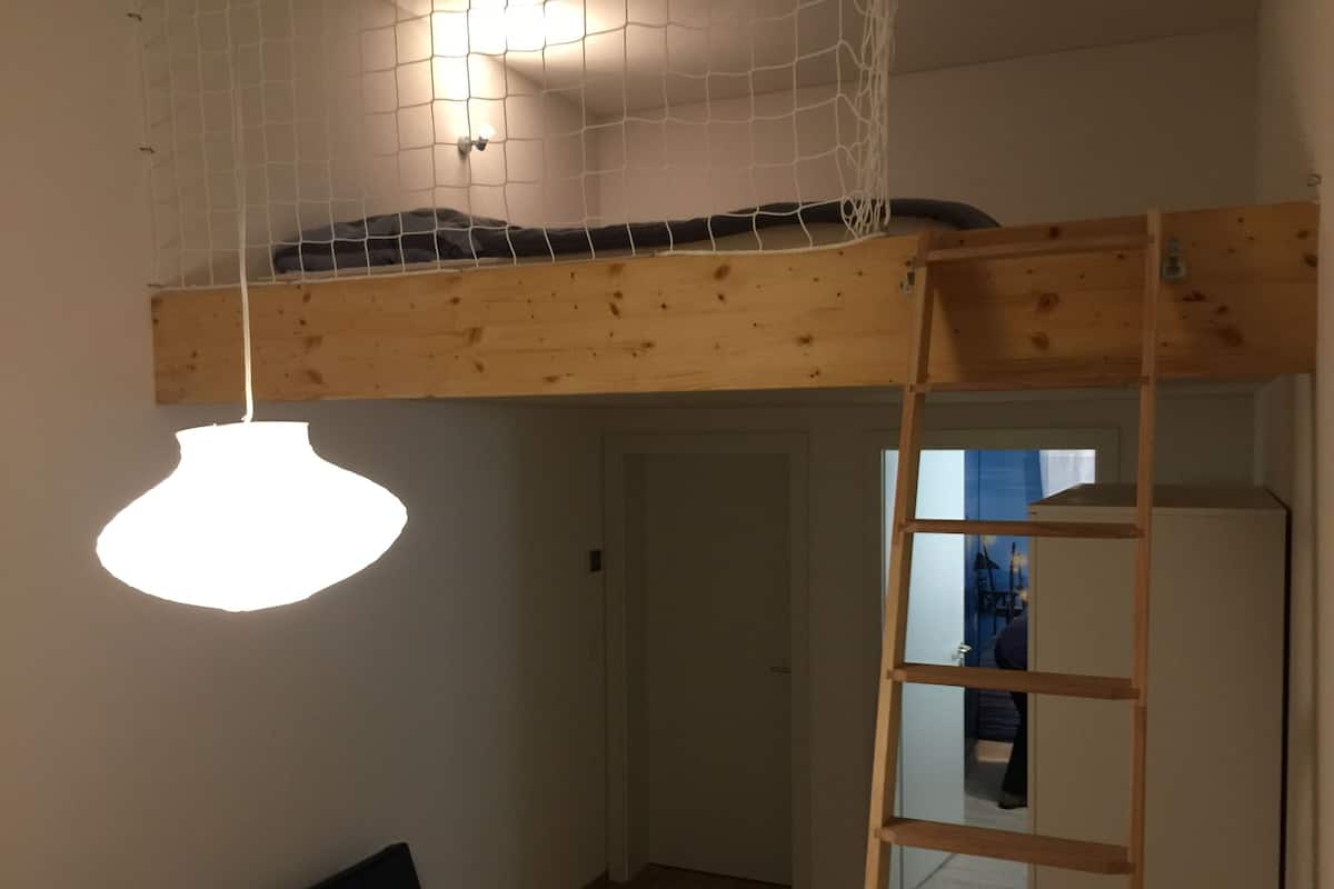 Une pièce avec un lit en mezzanine, une échelle, un luminaire suspendu et une porte donnant sur une autre pièce.