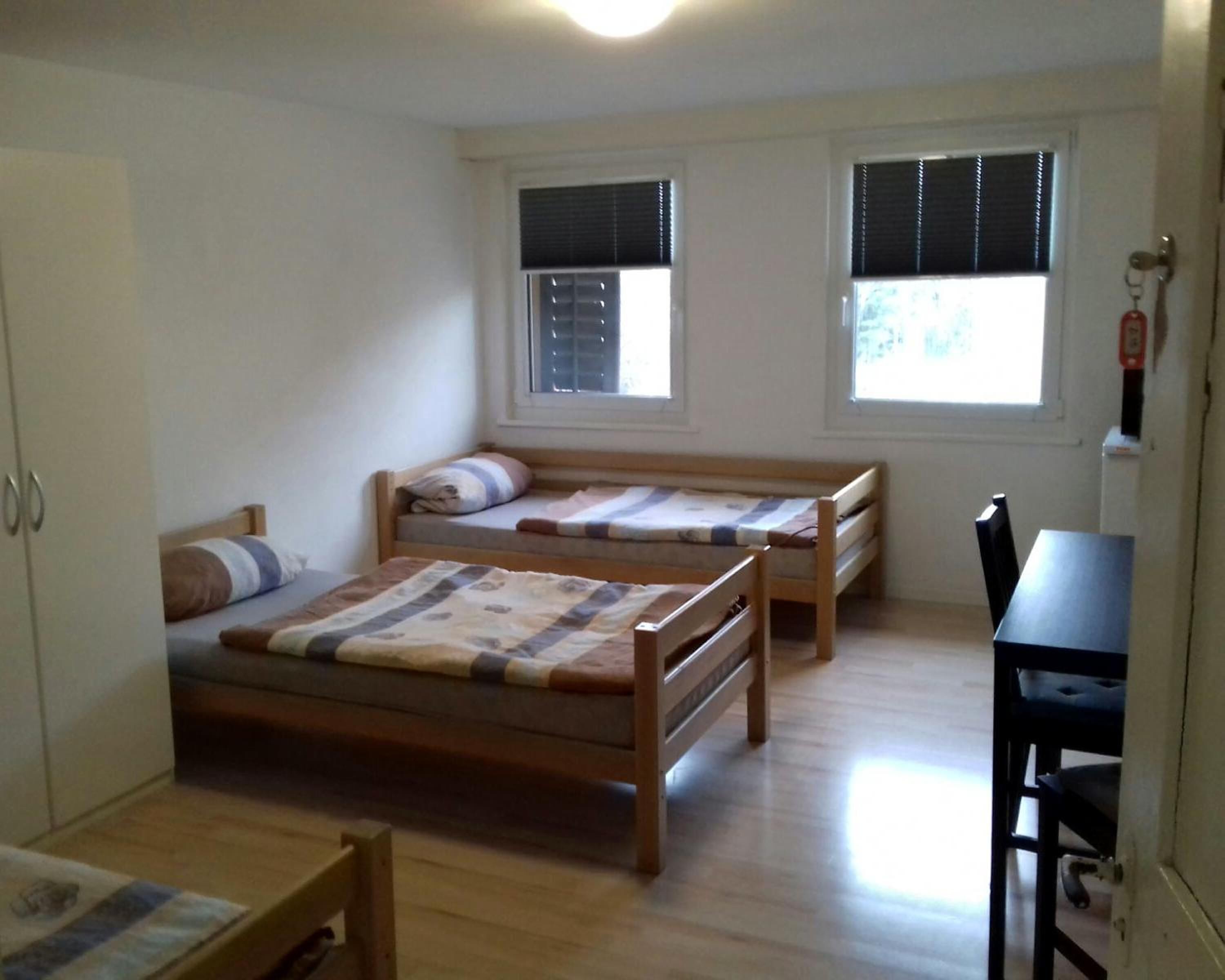 Una habitación pequeña y limpia, con dos camas de madera, un escritorio y dos ventanas con persianas.