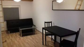 Une pièce avec un canapé noir, une table à manger avec des chaises, un miroir au mur et un plancher en bois.