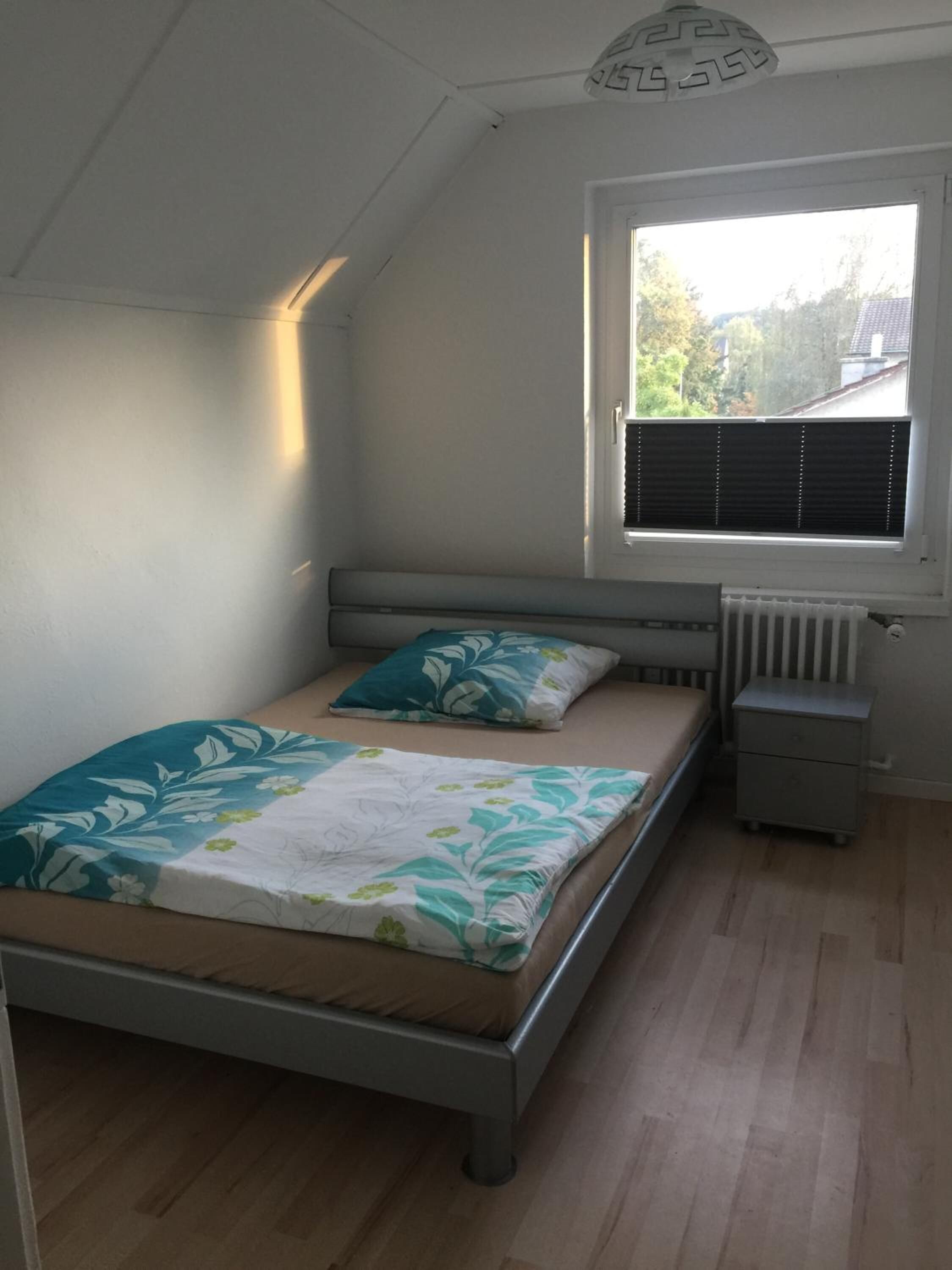 A bedroom with a bed, a nightstand, a window with blinds, and a radiator.
