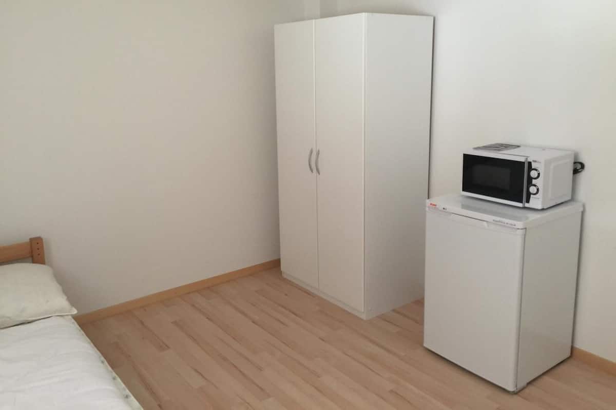 Une petite chambre propre, avec un lit, une armoire et un micro-ondes posé sur un mini-réfrigérateur.
