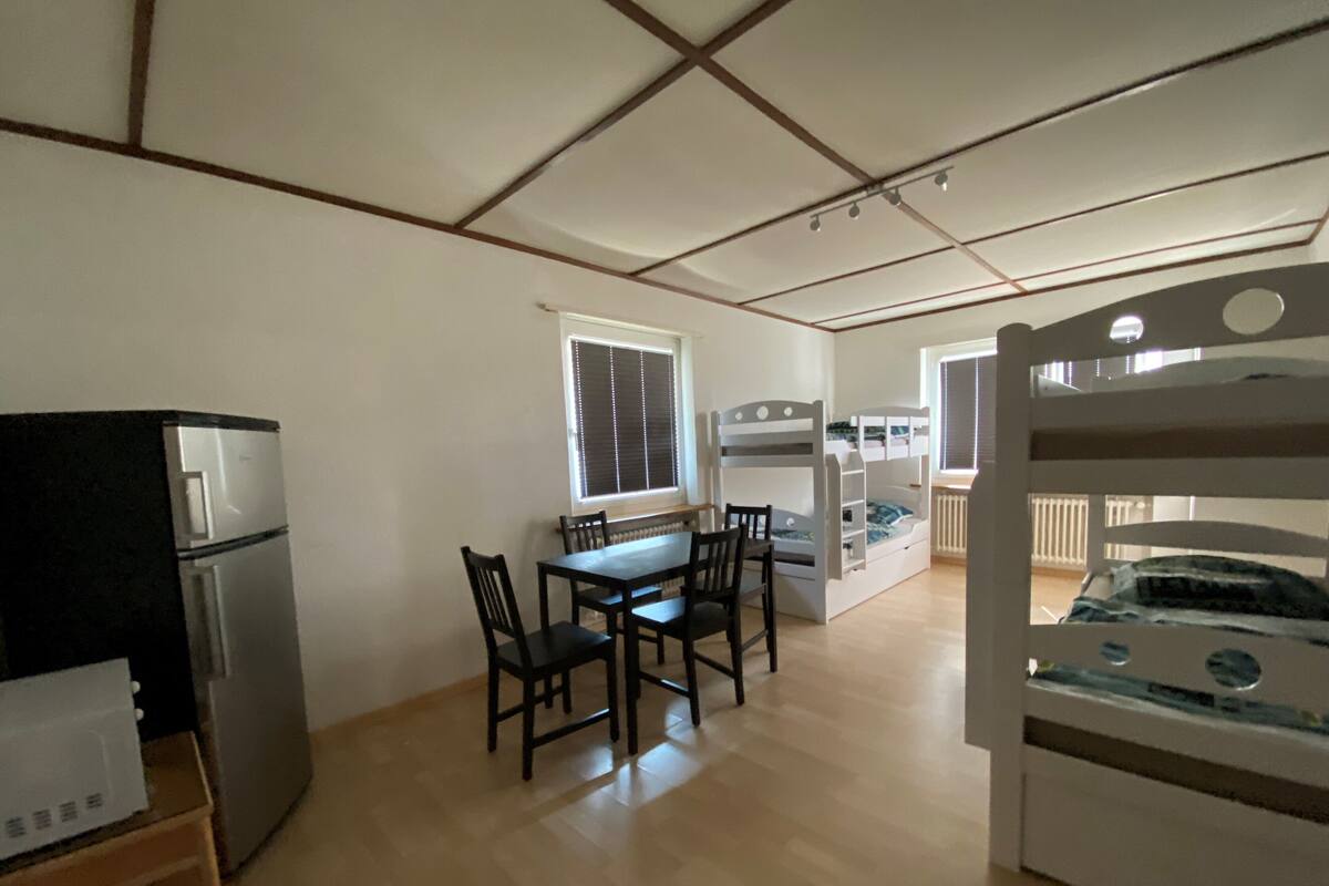 Une pièce compacte comprenant un coin cuisine, une table à manger avec des chaises, un lit superposé et une fenêtre avec des stores.