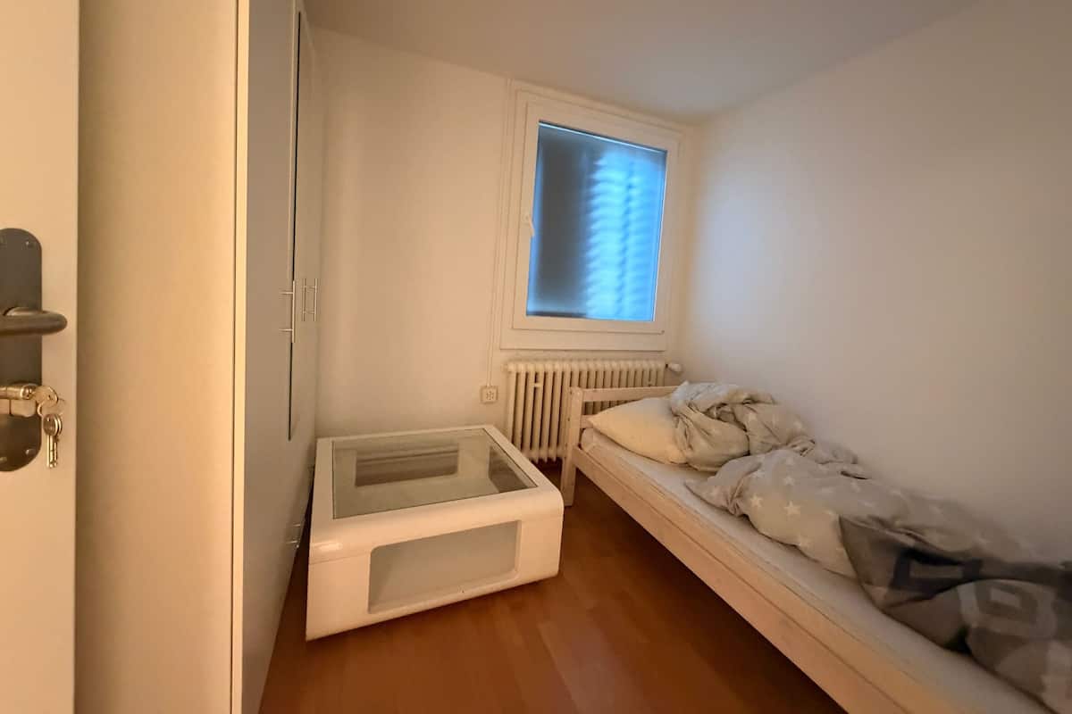 Une petite pièce, de taille modeste, comprenant un lit simple, un radiateur, une fenêtre avec des stores et une armoire blanche.