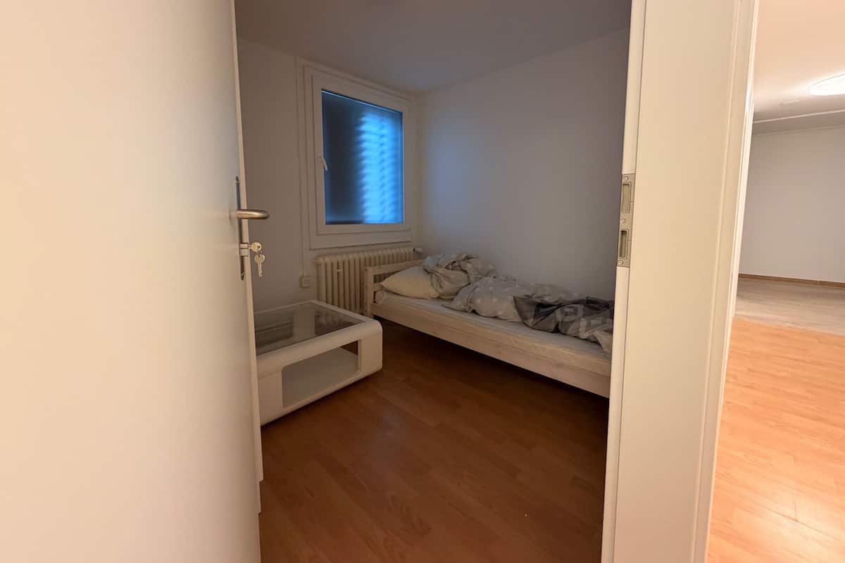 Un petit appartement d’une seule pièce, avec un plancher en bois, une fenêtre avec des stores, un radiateur et un lit recouvert d’une couverture à motifs.