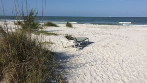 Una playa con una única silla de descanso blanca y una mesita, rodeada de arena y vegetación escasa.