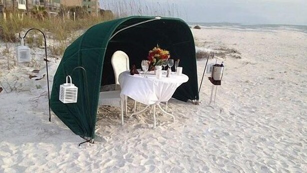 Una carpa de playa con una mesa preparada para dos, con mantel blanco y un jarrón de flores.