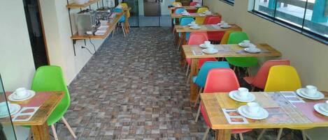 Un comedor con sillas y mesas de colores, piso de baldosas y vista a través de un ventanal.