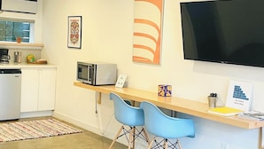 Una cocina moderna con armarios blancos, microondas y cafetera. Hay un televisor de pantalla plana grande montado en la pared y un escritorio de madera con dos sillas azules.