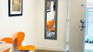 Una habitación con un escritorio, dos sillas anaranjadas, un cuadro enmarco, un espejo y una puerta que da al exterior.