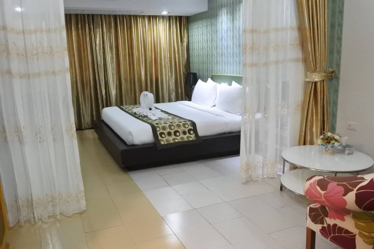 A hotel room with a bed, curtains, a small round table with a vase, and a chair.