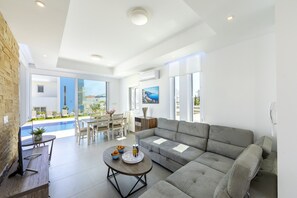 Un soggiorno moderno con un divano grigio, un tavolino e un'area pranzo con tavolo e sedie. Una grande finestra offre vista su una piscina e mobili da esterno.