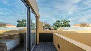 A modern balcony with a glass door, wooden flooring, and a view of surrounding buildings and trees.