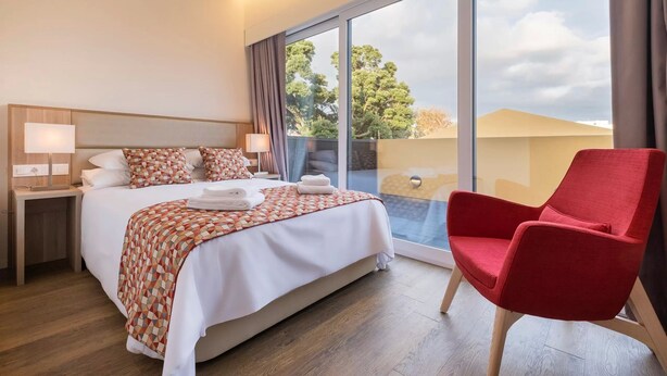 A hotel room with a large bed, a red armchair, a nightstand with a lamp, and a view of the outdoors through a large window.