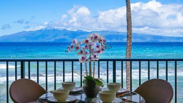 Mesa de comedor con sillas y un florero, con vista a una playa de aguas azul cristalino y montañas distantes.