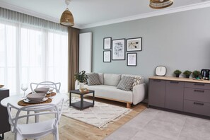 Een moderne woonkamer met een bank, eettafel en stoelen. Er hangen ingelijste foto's aan de muur, een koffieapparaat en een klok op het aanrecht.