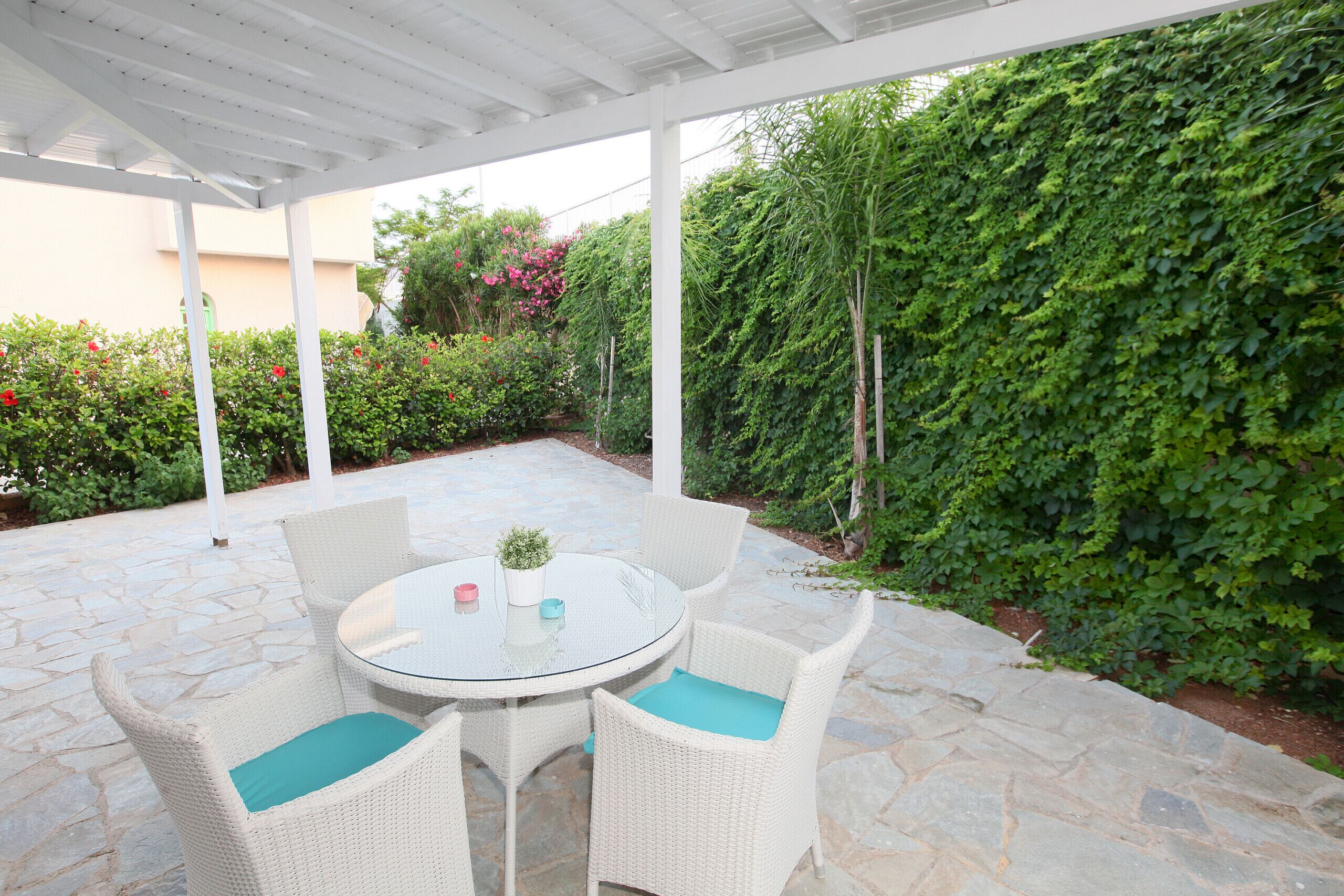 An outdoor patio with a round table and white wicker chairs, surrounded by stone flooring and lush greenery.