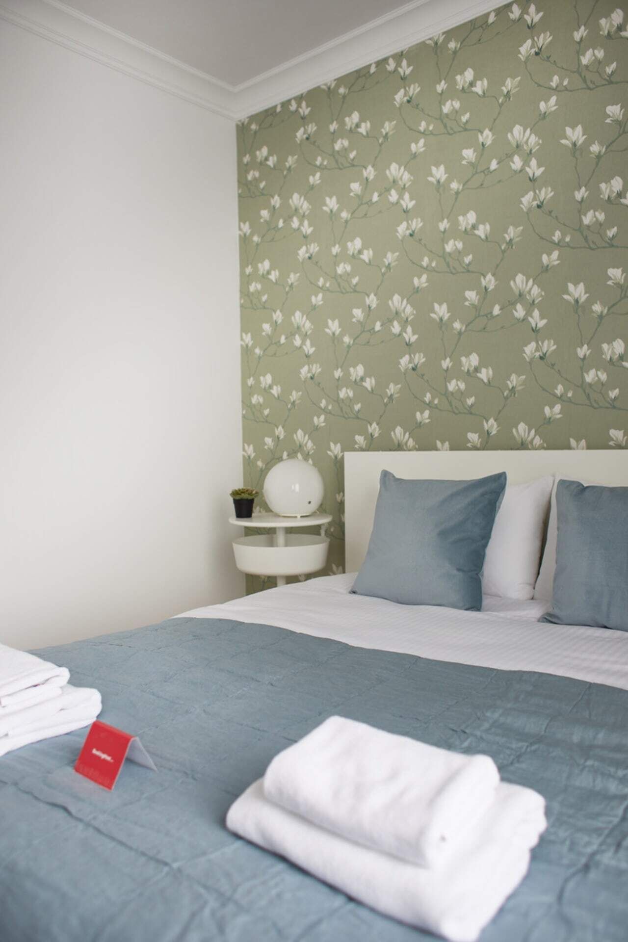 Una cama bien hecha con colcha y almohadas azules, una mesita de noche con una lámpara y una planta, y una pared con estampado de papel tapiz floral.
