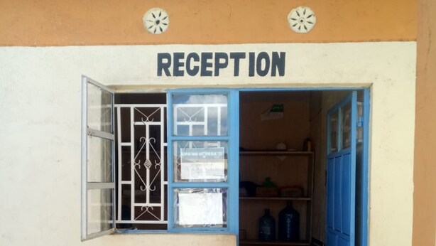 A reception area with a sign above the door, a window with a metal grill, a shelf with bottles, and a chair.