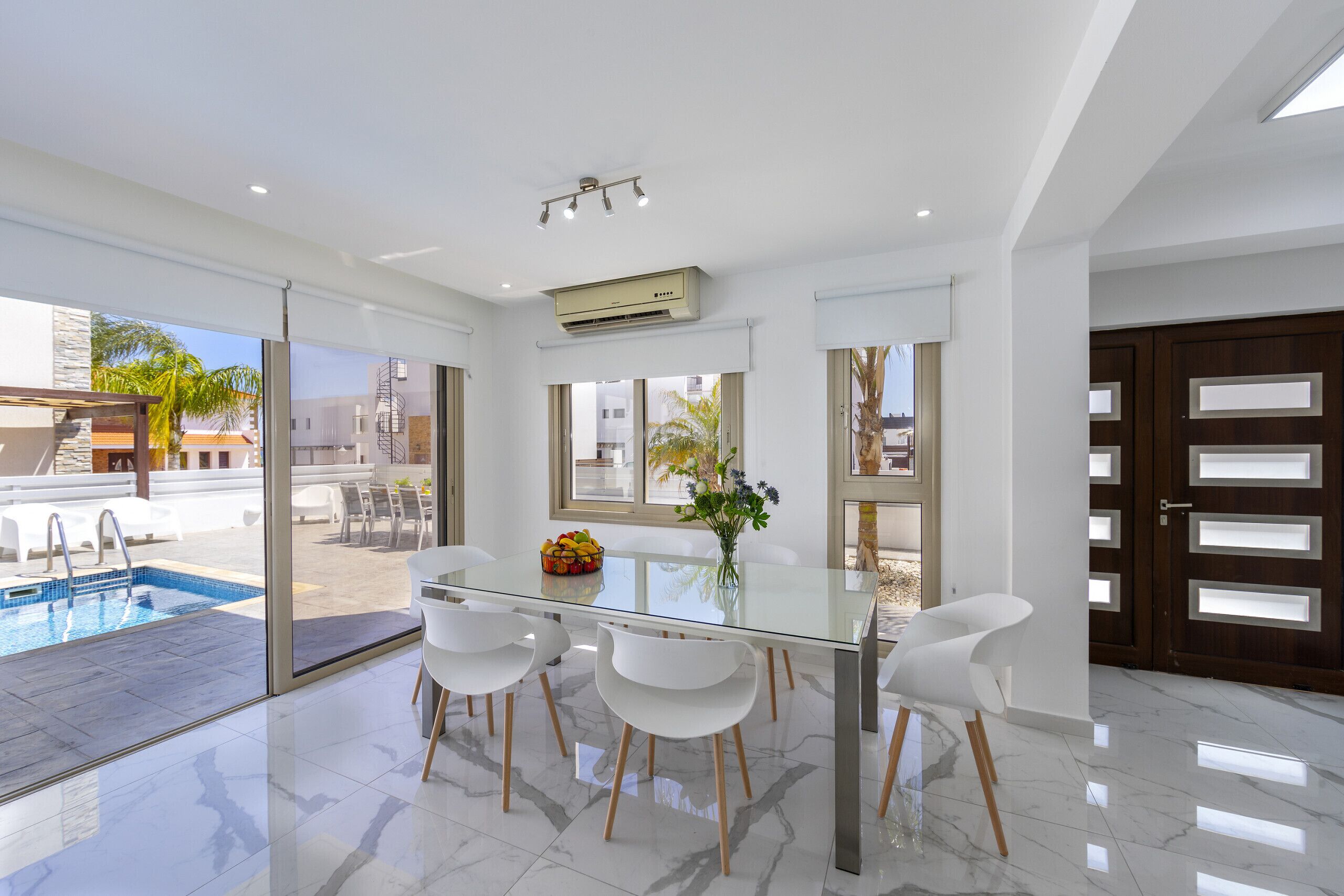 A modern dining area with a glass table, white chairs, and a view of a swimming pool.