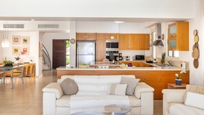 A modern kitchen with wooden cabinets, a central island, and stainless steel appliances. There is a dining area with a table and chairs, and a living room with a white sofa. - Protaras Beachfront Villa Pietra (Protaras)