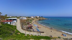A coastal beach with a pathway leading to a building with a red roof, a row of beach chairs, and a lifeguard tower. - Protaras Beachfront Villa Pietra (Protaras)