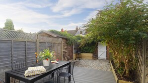 Une terrasse en bois avec une table et des chaises, entourée d’une clôture en bois et d’un jardin agrémenté de plantes et d’un petit arbre.