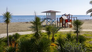 A lifeguard tower and playground on a beach with palm trees and a clear sky. - Mackenzie Castella Beachfront Suite (Larnaca)