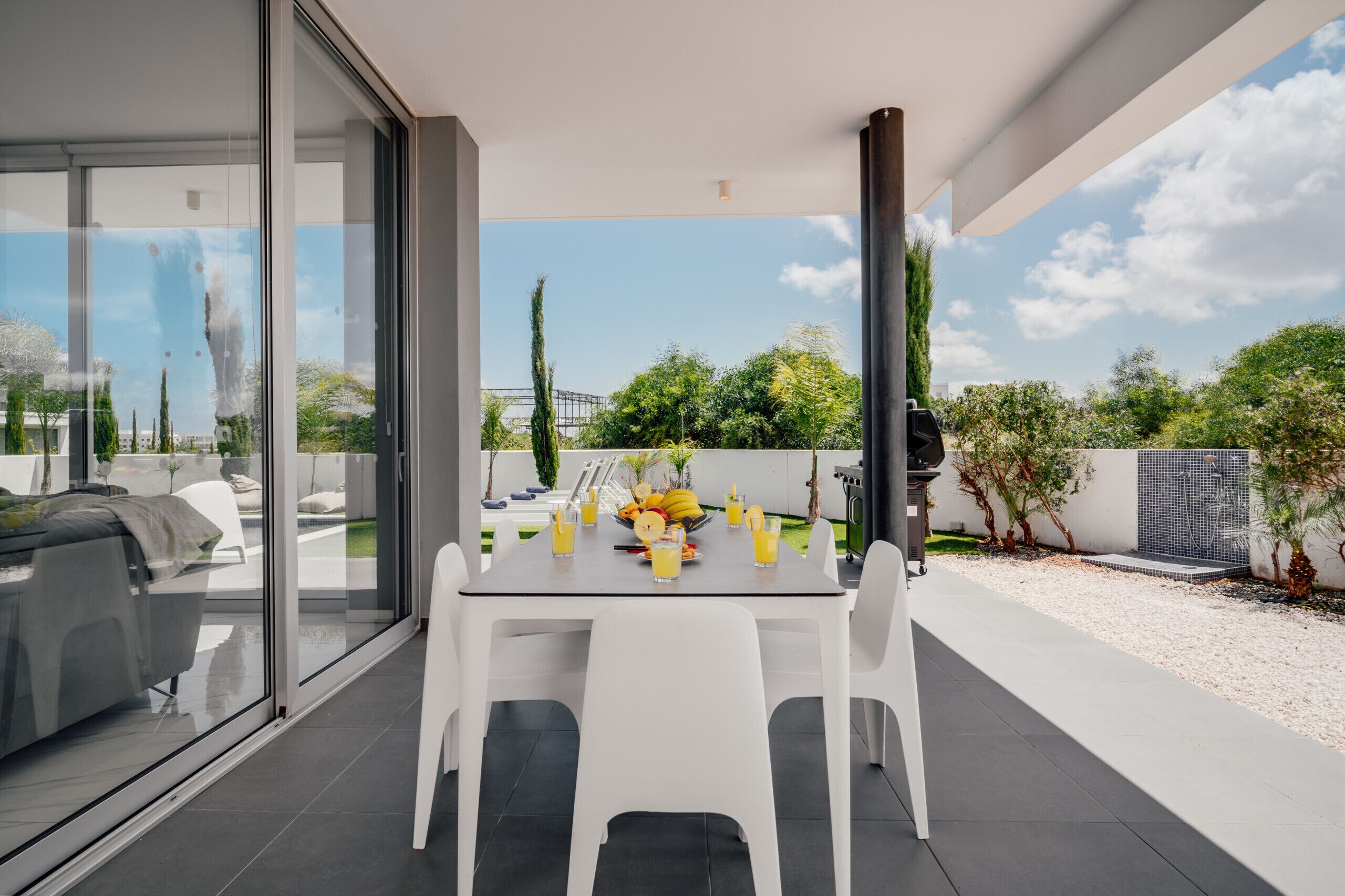 Een moderne eethoek buiten met een glazen wand, een witte tafel en stoelen, met uitzicht op een tuin en een heldere lucht.