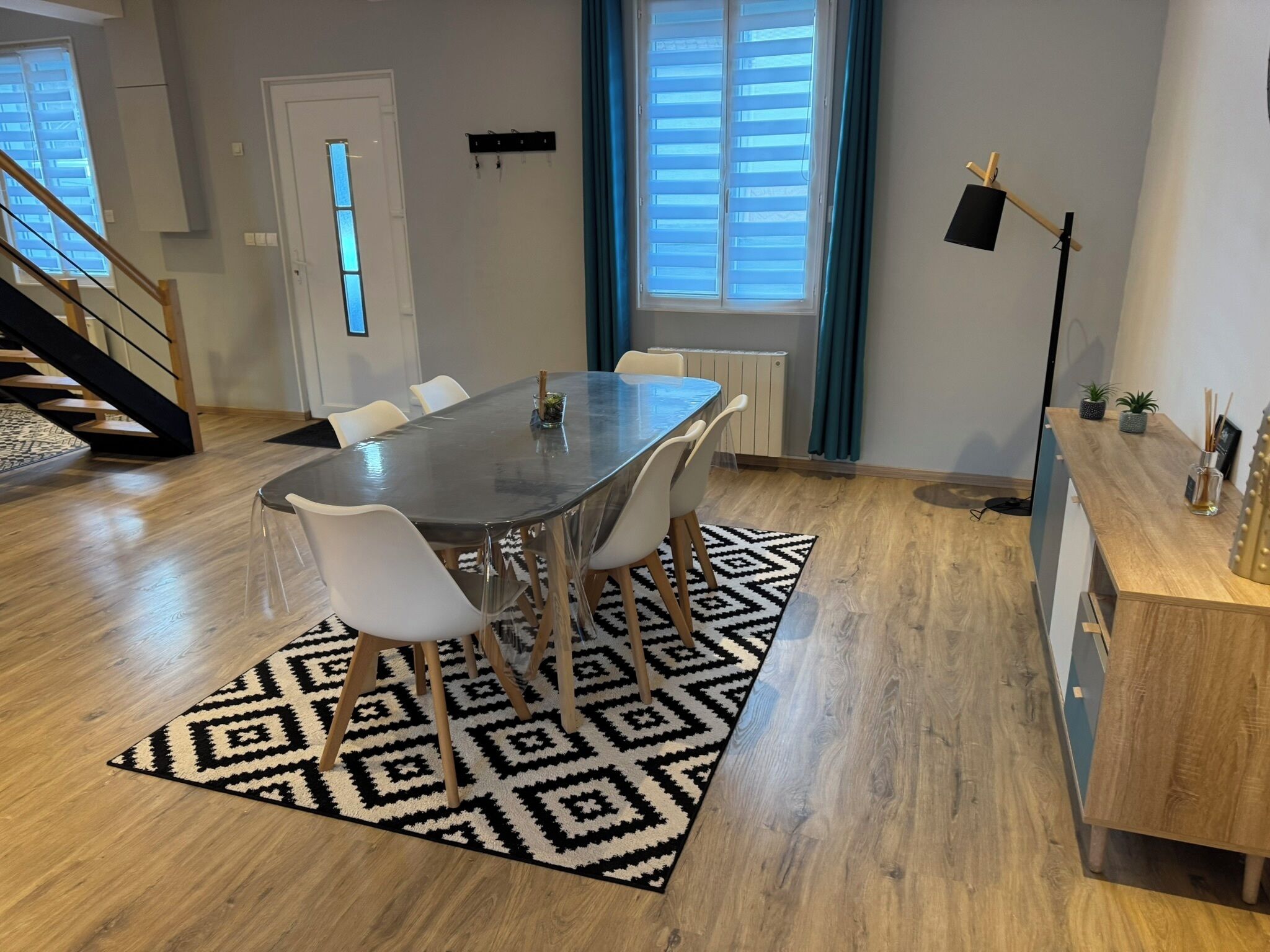 Un espace repas moderne avec une table rectangulaire, des chaises blanches, un tapis à motifs et un escalier en arrière-plan.