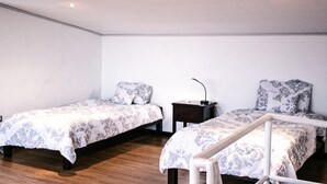 A room with two single beds, each with a patterned bedspread and matching pillows. There is a small bedside table with a lamp on it. The room has wooden flooring and white walls.