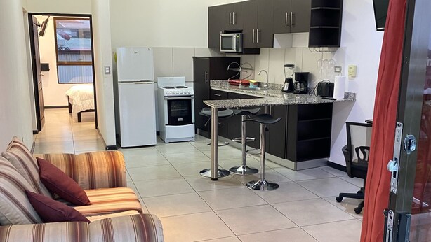 A modern kitchen with dark cabinets, a central island, and a ceiling fan. A dining area with a striped sofa and a red curtain is visible.