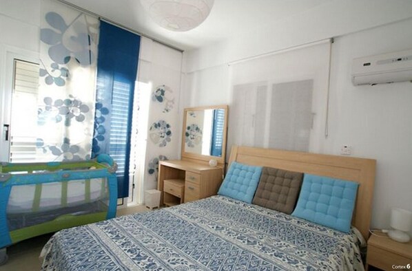 Een slaapkamer met een bed, een bureau, een stoel, een raam met zonwering en een wandairconditioner.