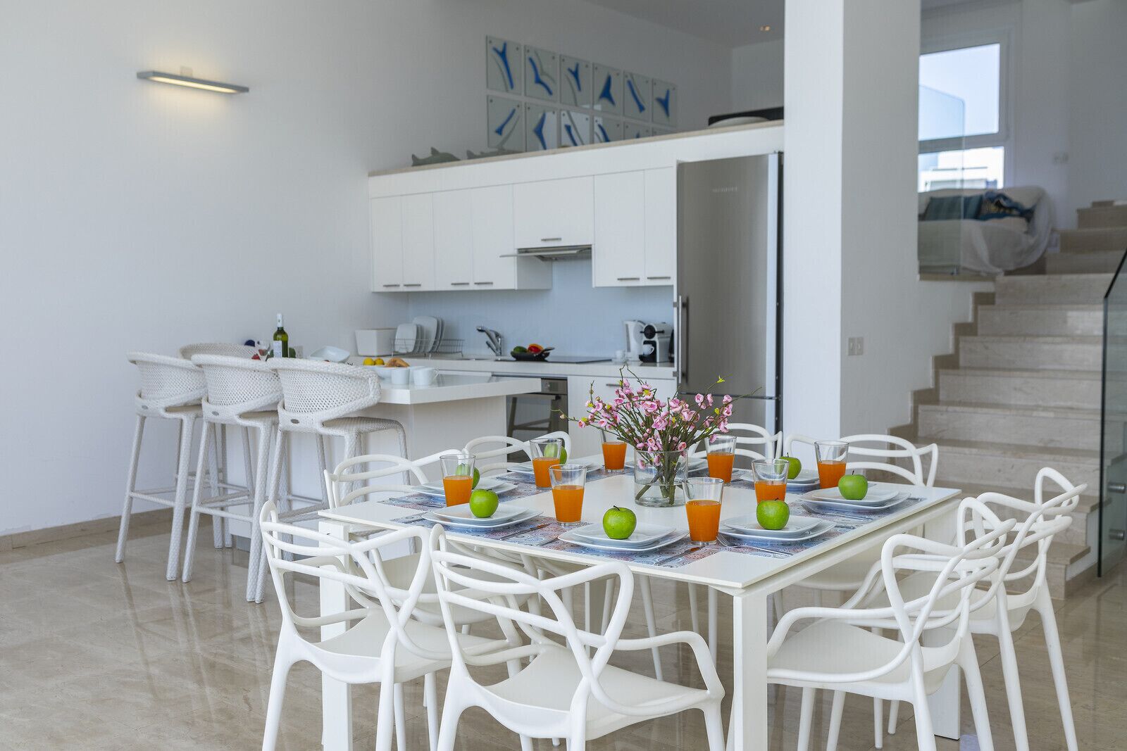 A modern kitchen with white cabinets, a dining area with white chairs, and a staircase leading to an upper level.