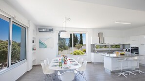 A modern kitchen with white cabinetry, a central island, and a dining area with white chairs. - Protaras Seaview Villa Chloe (Protaras)