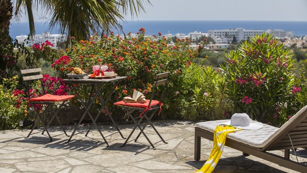 A patio with a table, chairs, a lounge chair, and a view of the sea and buildings. - Protaras Liora Seaview Suite (Protaras)