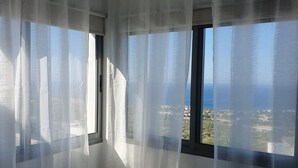 A bedroom with a bed, a window with sheer curtains, and a view of the sea and buildings. - Villa Imogine with Sea View (Neo Chorio)