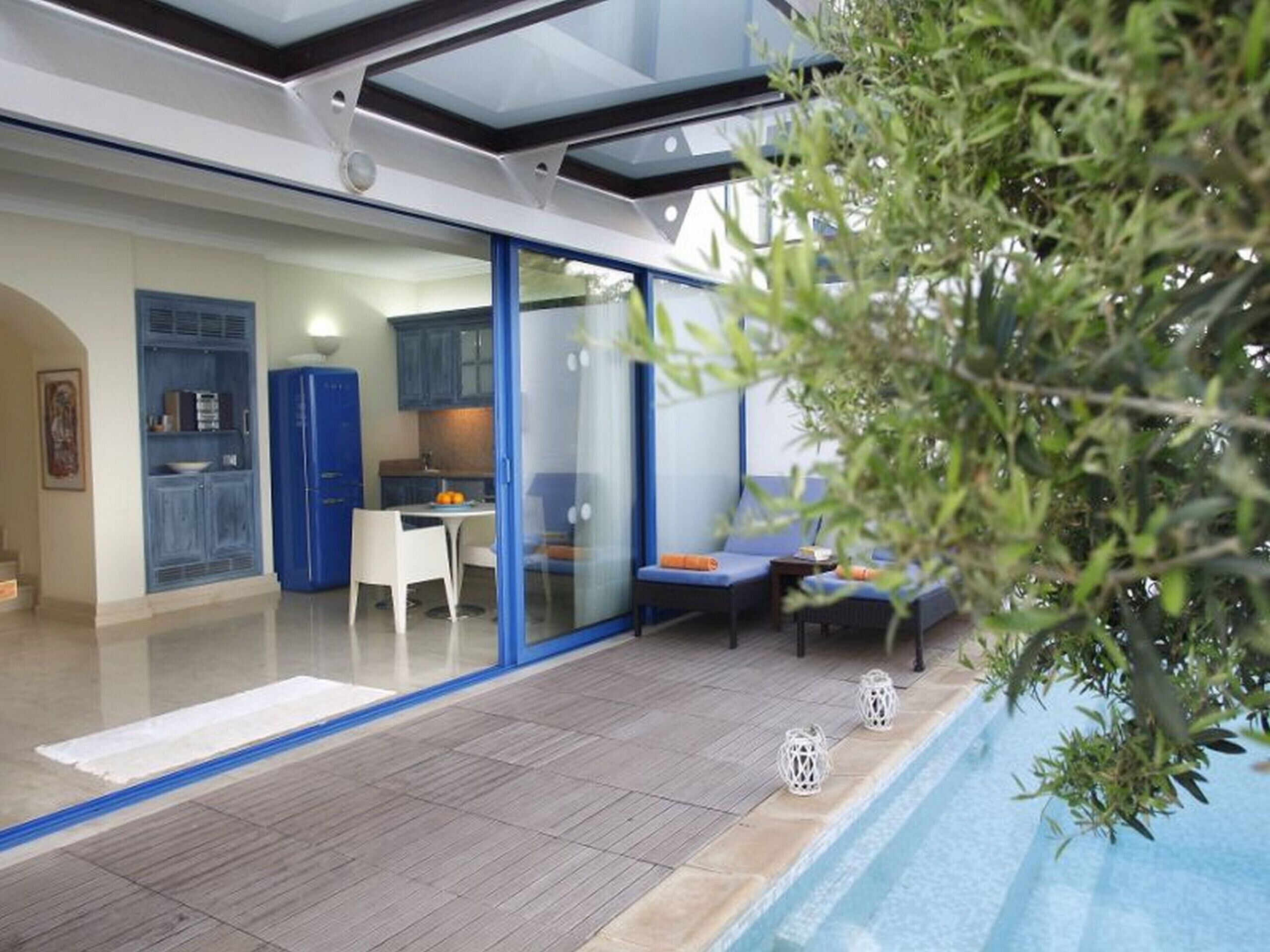 A modern indoor pool area with a glass-enclosed patio, a dining area, and a kitchen visible through the glass doors.