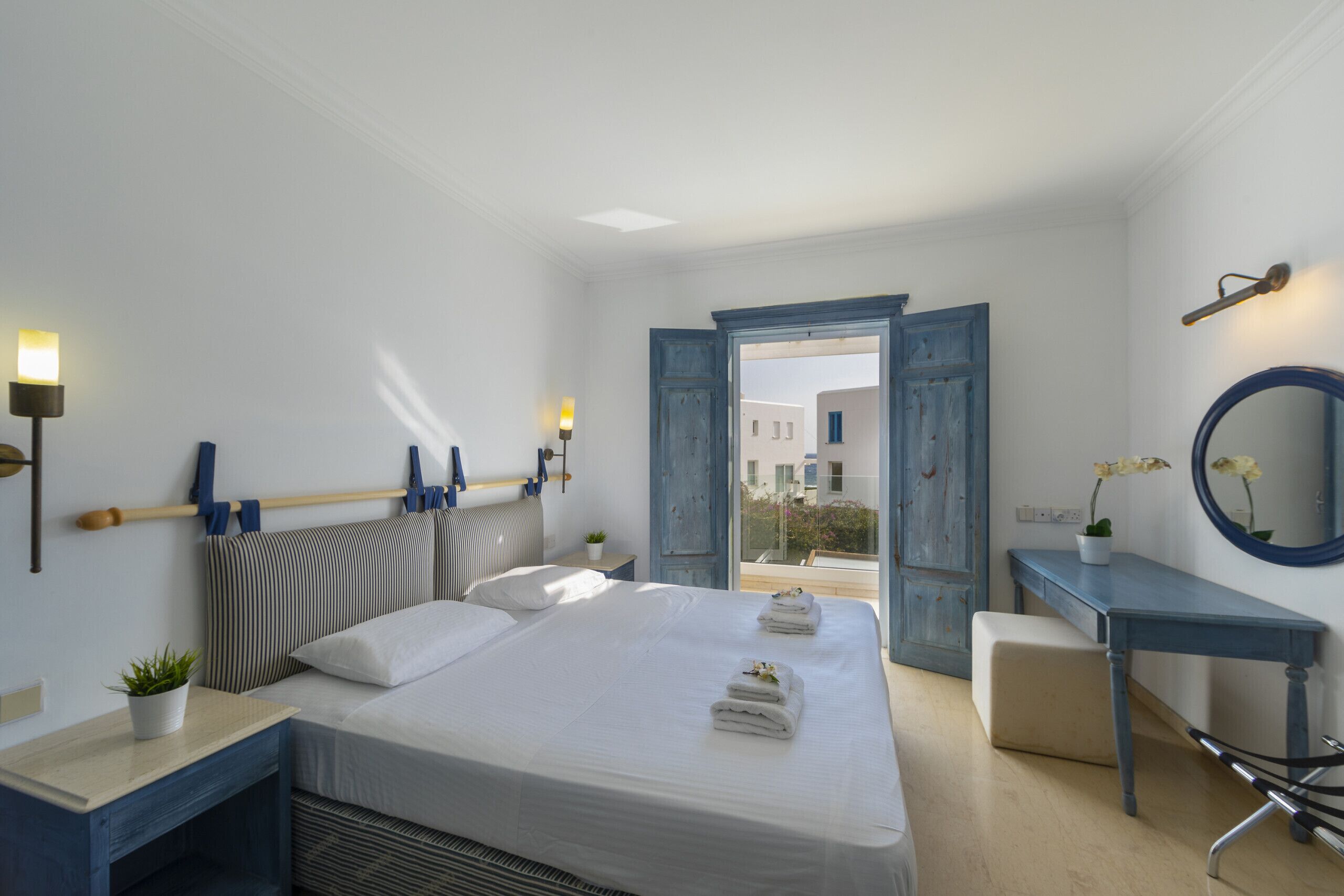 A bedroom with a bed, bedside tables, a desk, a mirror, and a view of the outside.