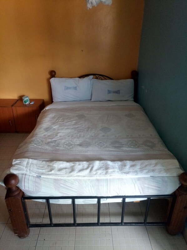 A single bed with a wooden frame and white bedding in a room with a tiled floor and a plain wall.