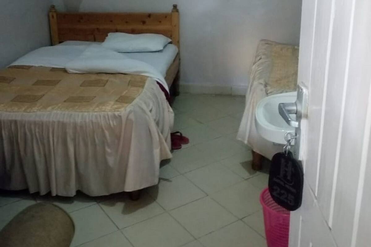 A small, simple room with a bed, a bedside table, a chair, a sink, and a hanging mosquito net.