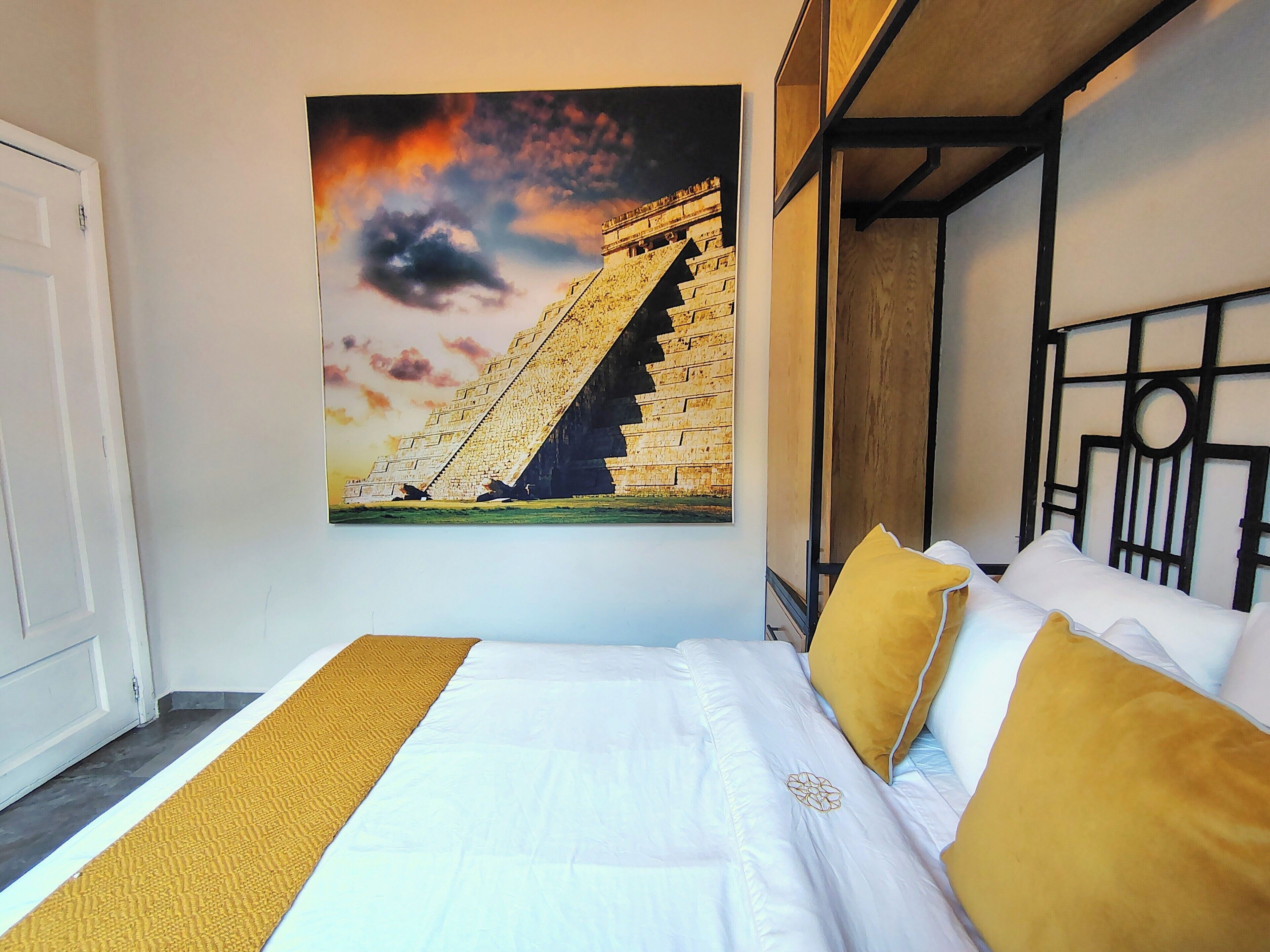 A hotel room with a bed, a headboard, a wall-mounted painting of a pyramid, and a wooden headboard frame.