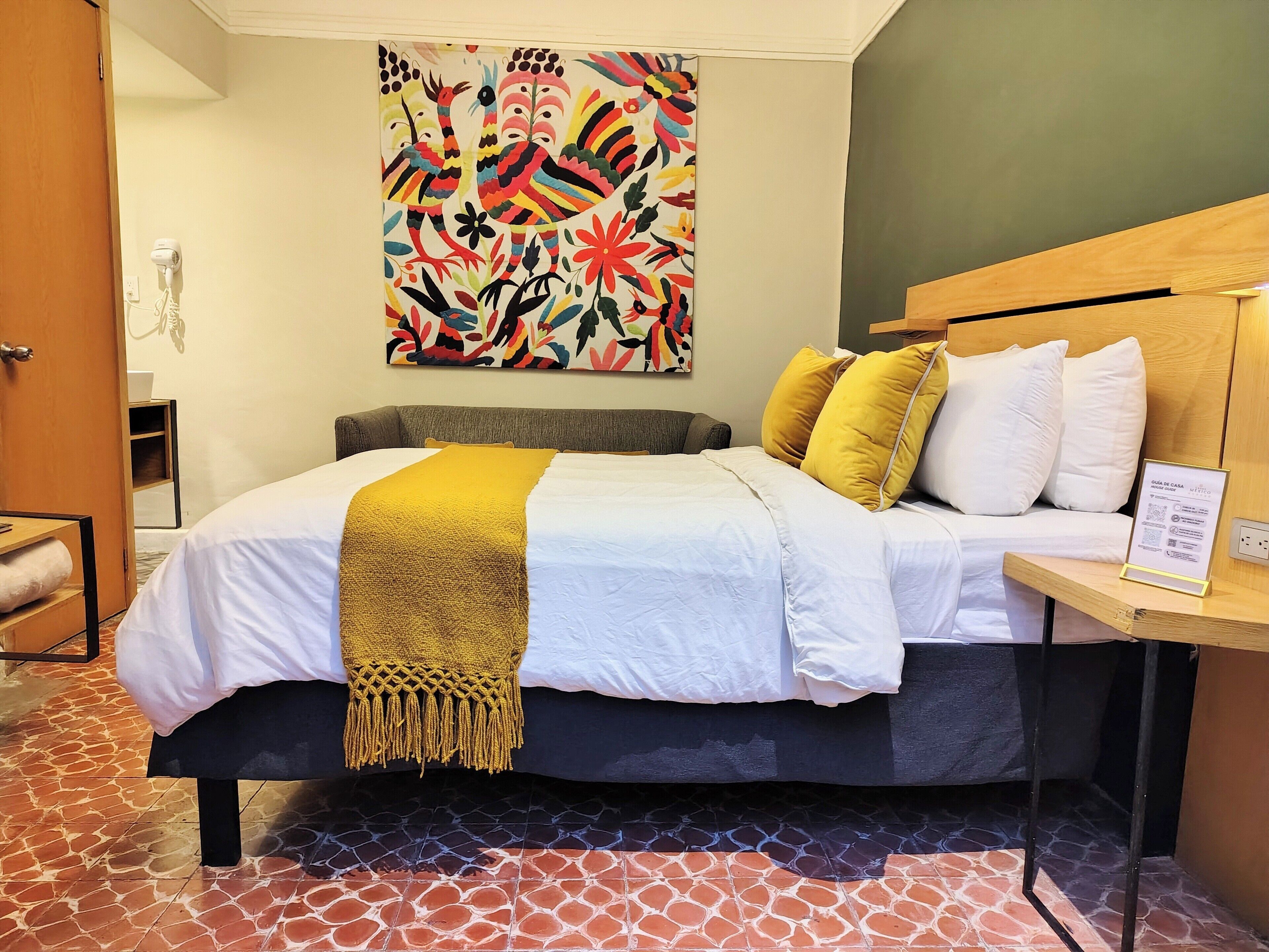 A hotel room with a bed, a headboard, a bedside table, a wall art piece, and a tiled floor.