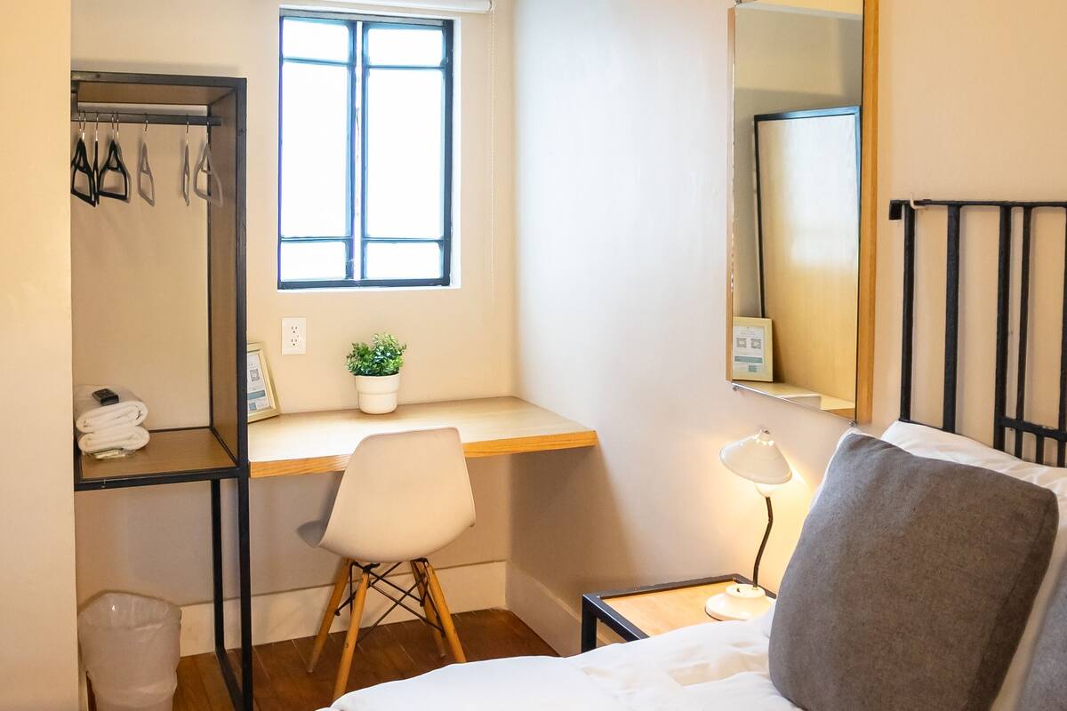Un dormitorio con cama, escritorio, silla, espejo y ventana.