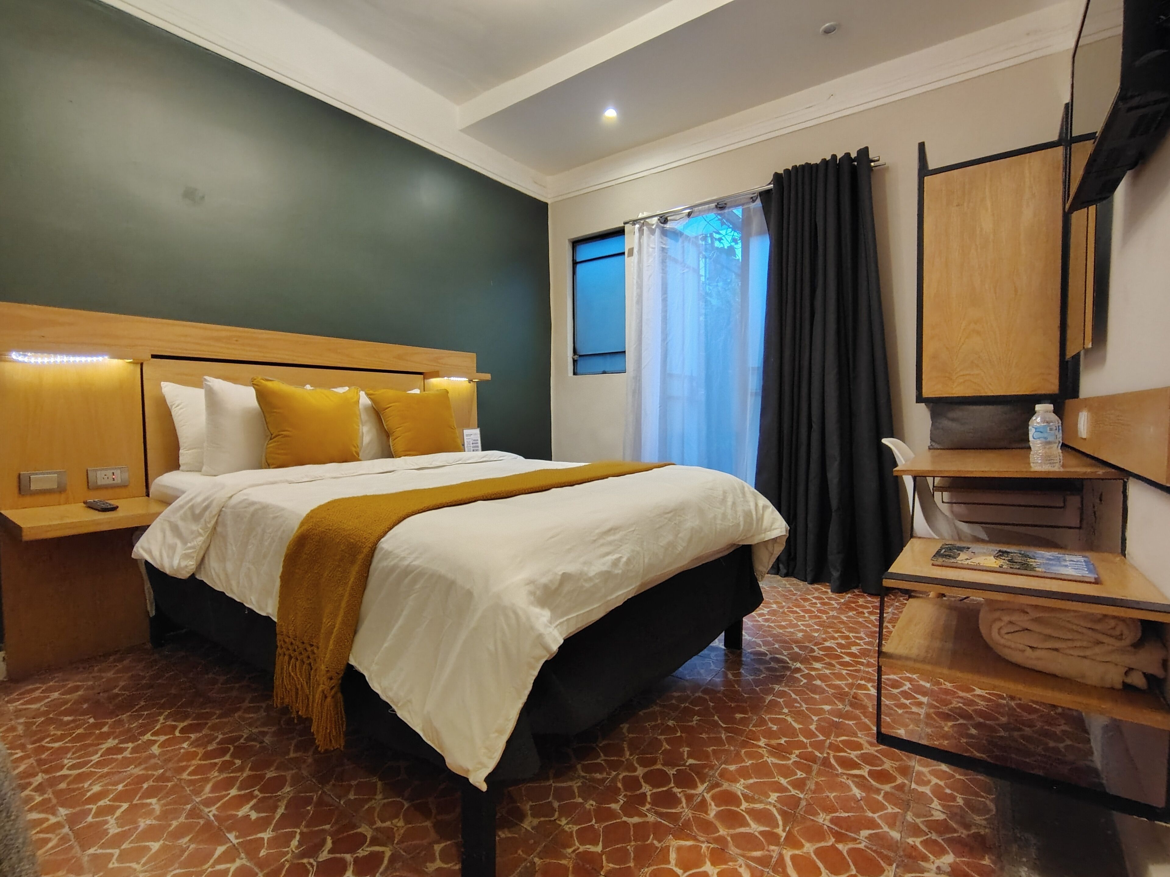 A hotel room with a bed, a nightstand, a wooden headboard, a mirror, a shelf with a water bottle, and a towel.
