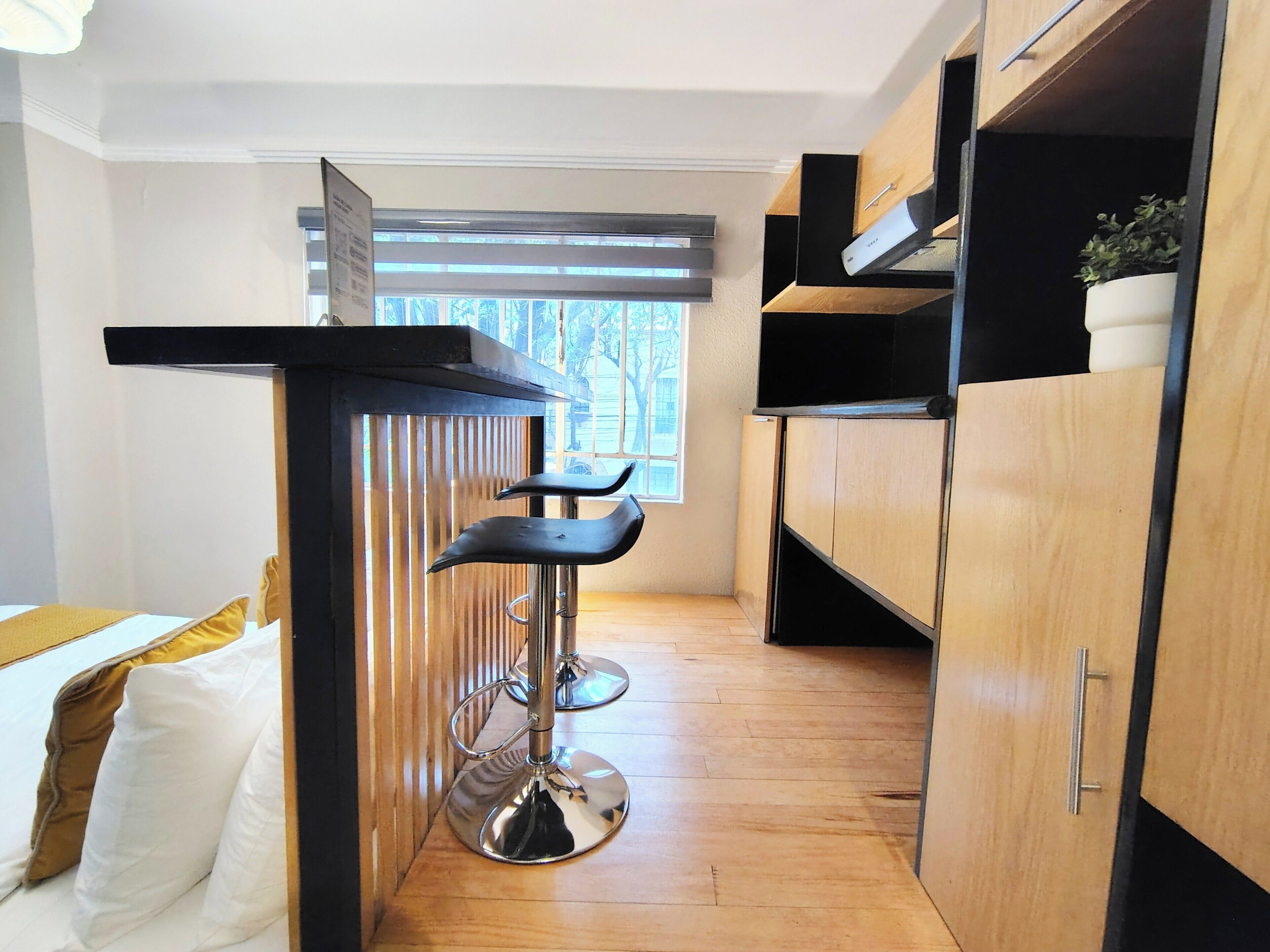 Habitación de hotel compacta con piso de madera, cama con ropa de cama blanca y amarilla, una ventana pequeña con persianas y una zona de kitchenette con cubierta y gabinetes negros.