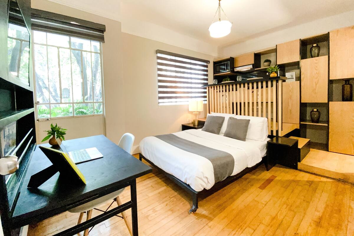 Un dormitorio con cama, un escritorio con computadora portátil, una silla, una ventana con persianas, un ventilador de techo y estantes empotrados.