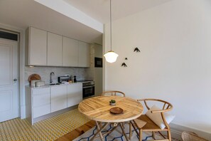 Une cuisine de taille réduite avec des meubles blancs, une table de salle à manger ronde en bois et un luminaire suspendu.