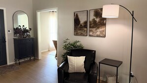 Een zithoek met een zwarte lederen fauteuil, een vloerlamp en een bijzettafeltje. Er hangt een spiegel aan de muur en een deur die naar een andere kamer leidt.