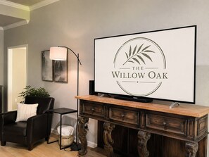 A living room with a flat-screen TV on a wooden console, a black leather chair, a floor lamp, and a framed picture on the wall.
