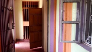 Um corredor estreito com uma porta de madeira no fim, um portão com grades à esquerda e uma janela à direita.